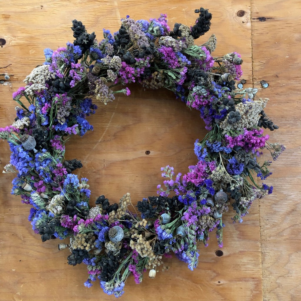 A handmade wreath made of various dried flowers in shades of purple, blue, and beige, placed on a wooden surface.